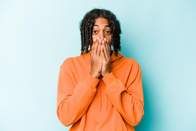 Junger afroamerikanischer Mann isoliert auf blauem Hintergrund schockiert, der den Mund mit den Händen bedeckt