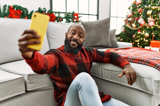 Junger afroamerikanischer Mann macht ein Selfie mit dem Smartphone, während er zu Hause am Weihnachtsbaum sitzt