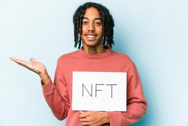 Junger afroamerikanischer Mann mit NFT-Plakat isoliert auf blauem Hintergrund, der einen Kopierraum auf einer Handfläche zeigt und eine andere Hand an der Taille hält