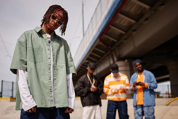 Junger afroamerikanischer Mann mit Sonnenbrille und stilvoller grauer Jacke steht gegen eine Gruppe interkultureller Freunde und schaut in die Kamera