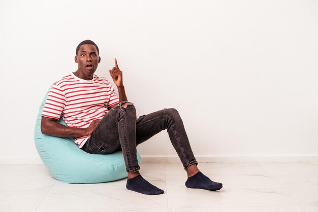 Junger afroamerikanischer Mann sitzt auf einem Puff isoliert auf weißem Hintergrund und hat eine Idee, ein Inspirationskonzept.
