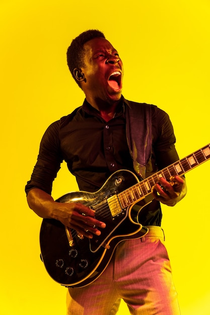 Junger afroamerikanischer Musiker, der Gitarre wie ein Rockstar auf gelbem Hintergrund im Neonlicht spielt. Konzept von Musik, Hobby, Festival, Open-Air. Fröhlicher Kerl improvisiert, singt Lied.