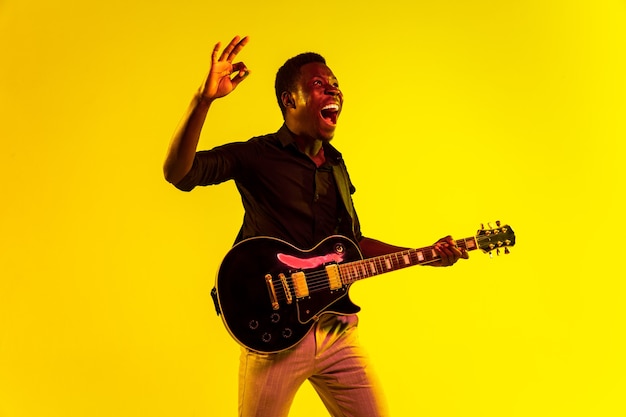 Junger afroamerikanischer Musiker, der Gitarre wie ein Rockstar auf gelbem Hintergrund im Neonlicht spielt. Konzept von Musik, Hobby, Festival, Open-Air. Fröhlicher Kerl improvisiert, singt Lied.