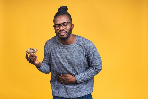 Junger afroamerikanischer schwarzer Mann mit Afro-Haar, der Donut isoliert auf gelbem Hintergrund mit der Hand auf dem Bauch hält, schmerzhafte Krankheit, die sich unwohl fühlt. Ache-Konzept.