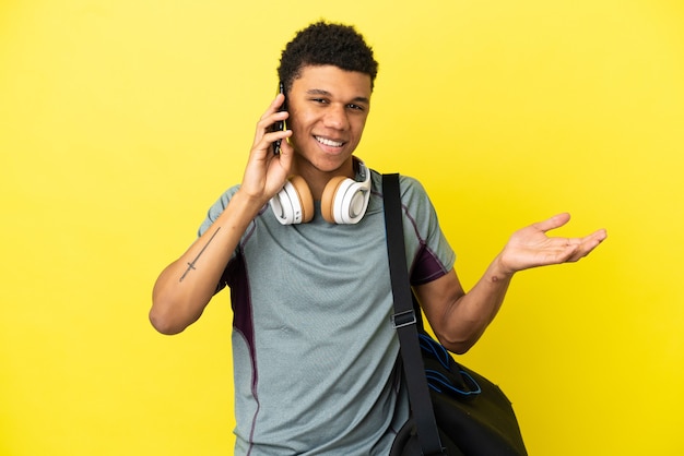Junger afroamerikanischer Sportmann mit Sporttasche einzeln auf gelbem Hintergrund, der ein Gespräch mit dem Handy mit jemandem führt