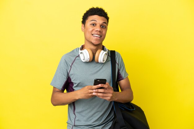 Junger afroamerikanischer Sportmann mit Sporttasche isoliert auf gelbem Hintergrund überrascht und sendet eine Nachricht