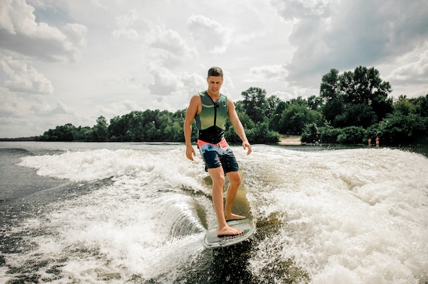 Junger aktiver Mann, der auf dem Brett hinunter den Fluss gegen den bewölkten Himmel und die Bäume wakesurfing ist