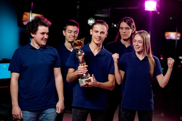 Junger Anführer des Cybersportteams hält goldenen Pokal, während er unter seinen Freunden mit glücklichem Mädchen nahe steht, indem er Triumph ausdrückt