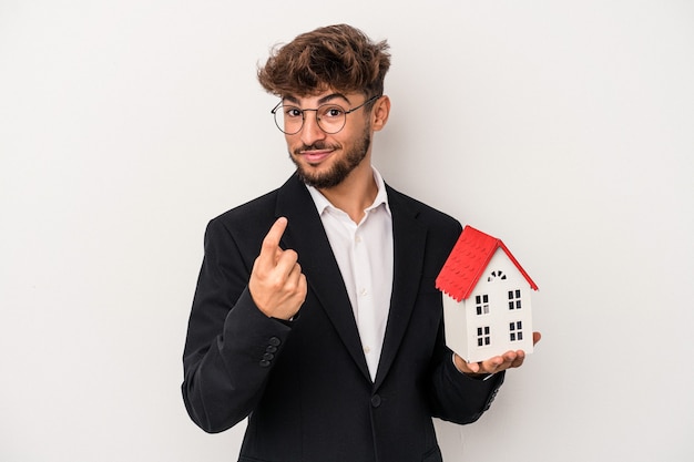 Junger arabischer Immobilienmann, der ein Musterhaus hält, das auf isoliertem Hintergrund isoliert ist und mit dem Finger auf Sie zeigt, als ob er einladen würde, näher zu kommen.