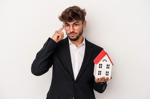 Junger arabischer Immobilienmann, der ein Musterhaus isoliert auf isoliertem Hintergrund hält und mit dem Finger auf den Tempel zeigt, denkt, konzentriert sich auf eine Aufgabe.