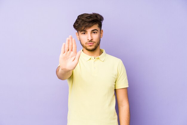 Junger arabischer Mann auf einer lila Wand, die mit ausgestreckter Hand steht, die Stoppschild zeigt