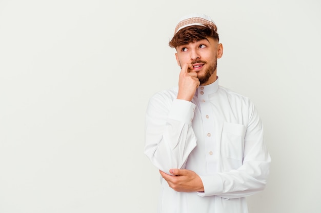Junger arabischer Mann, der das typische arabische Kostüm lokalisiert auf weißem entspanntem Denken über etwas betrachtet, das einen Kopienraum betrachtet.
