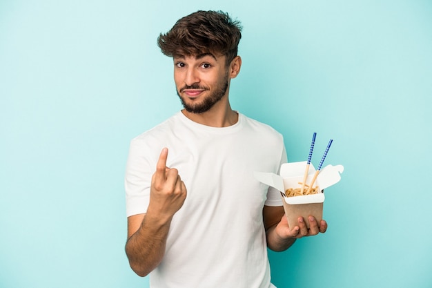 Junger arabischer mann, der eine nudel zum mitnehmen hält, die auf blauem hintergrund isoliert ist und mit dem finger auf sie zeigt, als ob sie einladen, näher zu kommen.