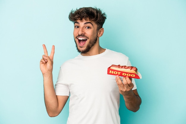 Junger arabischer Mann, der einen Hotdog isoliert auf blauem Hintergrund hält, freudig und sorglos, der ein Friedenssymbol mit den Fingern zeigt.