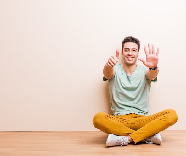 Junger arabischer Mann, der freundlich lächelt und schaut, Nummer sechs oder Sechste mit der Hand nach vorne zeigend, auf dem Boden sitzend herunterzählend