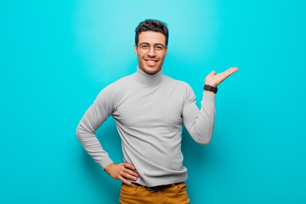 Junger arabischer Mann, der lächelt, sich sicher, erfolgreich und glücklich fühlt und Konzept oder Idee auf Kopienraum auf der Seite gegen flache Wand zeigt