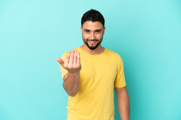 Junger arabischer Mann isoliert auf blauem Hintergrund, der einlädt, mit der Hand zu kommen. Schön, dass du gekommen bist