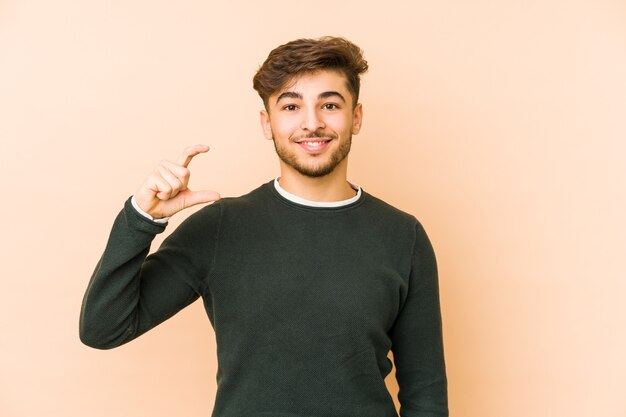 Junger arabischer Mann lokalisiert auf einer beigen Wand, die etwas wenig mit Zeigefingern hält, lächelnd und zuversichtlich.