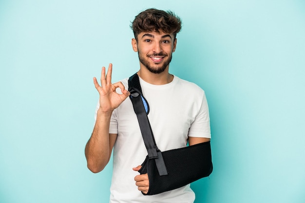 Junger arabischer mann mit gebrochener hand isoliert auf blauem hintergrund fröhlich und selbstbewusst, der eine ok geste zeigt.