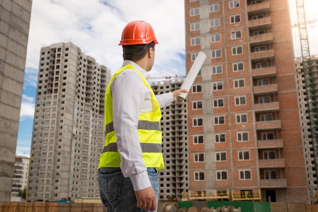 Junger Architekt in Bauarbeiterhelm und Warnweste, der Gebäude mit gerollten Blaupausen zeigt