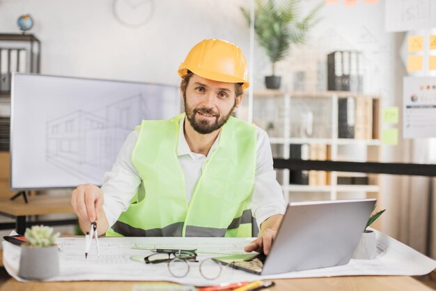 Foto junger architekt in helm und uniform sitzt mit bauplänen und modernen geräten am tisch