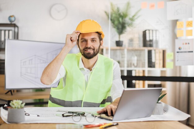 Junger Architekt in Helm und Uniform sitzt mit Bauplänen und modernen Geräten am Tisch