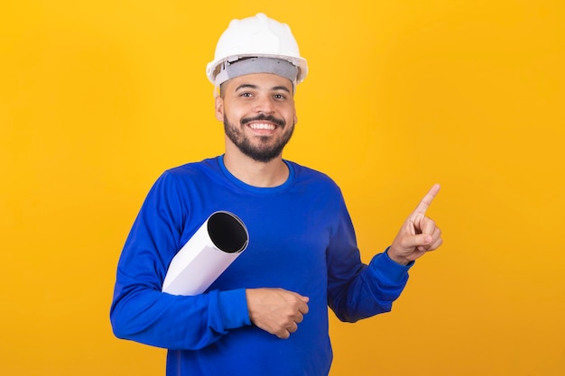 Junger Architekt mit Helm und Blaupausen isoliert auf gelbem Hintergrund, der mit dem Finger zur Seite zeigt