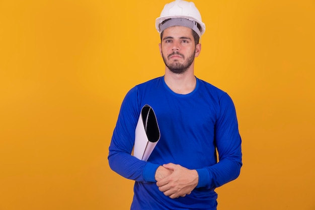 Junger architekt mit helm und blaupausen isoliert auf gelbem hintergrund, der mit dem finger zur seite zeigt