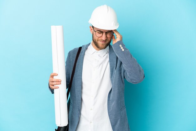 Junger Architektenmann mit Helm und hält Blaupausen über lokalisiertem Hintergrund mit Kopfschmerzen