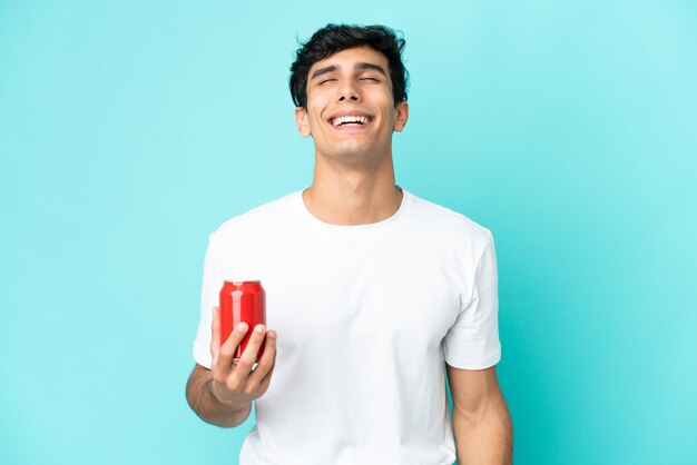 Junger argentinischer Mann, der eine Erfrischung isoliert auf blauem Hintergrund hält, lacht