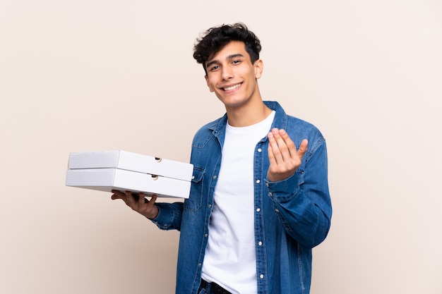 Junger argentinischer Mann, der Pizzas über der lokalisierten Wand einlädt, mit der Hand zu kommen hält. Schön, dass Sie gekommen sind