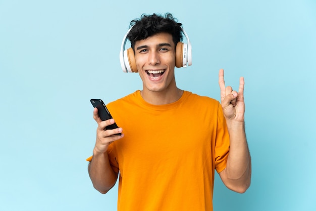 Junger argentinischer Mann lokalisiert an der Wand, die Musik mit einem Handy hört, das Rockgeste macht