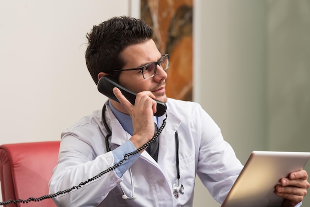 Junger Arzt, der an seinem Computer arbeitet, während er am Telefon spricht