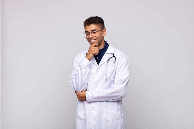 Junger Arzt Mann lächelnd mit einem glücklichen, selbstbewussten Ausdruck mit der Hand am Kinn