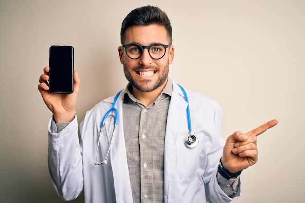 Junger Arzt mit Stethoskop zeigt Smartphone-Bildschirm vor isoliertem Hintergrund und zeigt sehr glücklich mit der Hand und dem Finger zur Seite