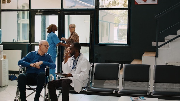 Foto junger arzt spricht mit älterem mann in der empfangslobby und führt medizinische beratung mit patienten mit chronischer behinderung durch. rollstuhlfahrer bitten mediziner um hilfe und behandlung zur heilung von krankheiten.