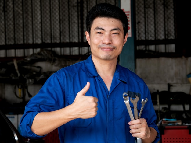 Junger asiatischer Automechaniker in der Uniform, die Schlüssel hält und Daumen oben in der Garage zeigt.
