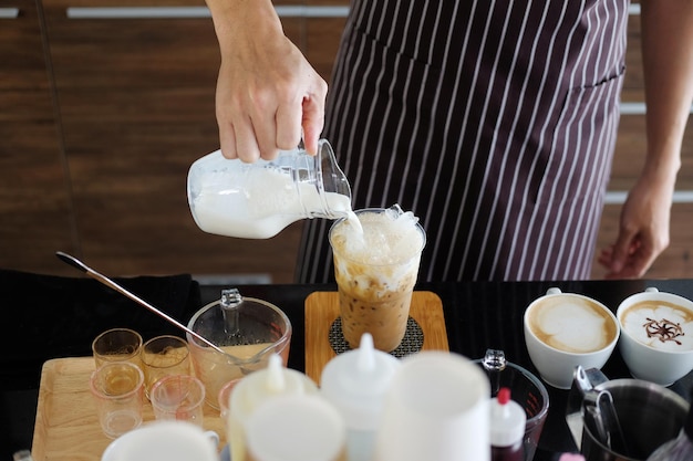 Junger asiatischer barista-mann, der frische milch in ein plastikglas latte-eiskaffee zur bestellung gießt