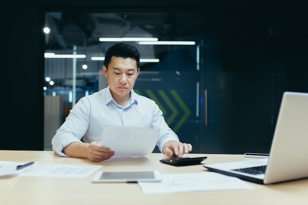 Junger asiatischer Buchhalter, der mit Dokumenten und Taschenrechner arbeitet und an einem Schreibtisch mit Laptop sitzt