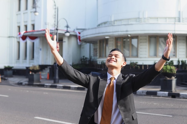 junger asiatischer Geschäftsmann, der glücklich ist und die Hände hebt, während er im Freien mit einem Bürogebäude steht