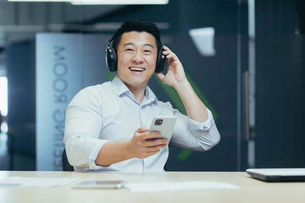 Junger asiatischer geschäftsmann hört musik über kopfhörer und machte am telefon eine pause von der arbeit