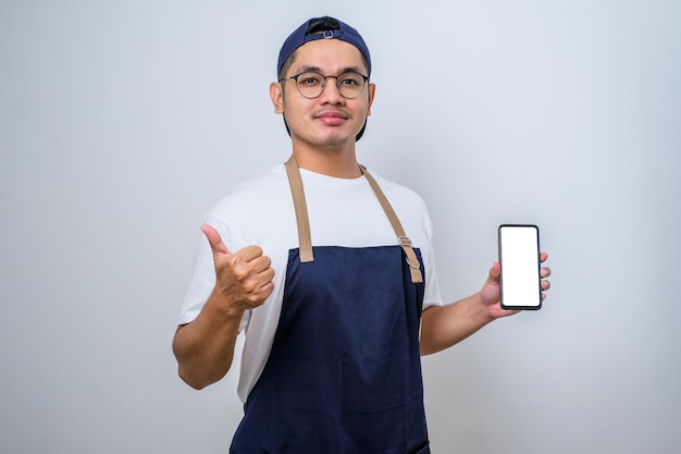 Junger asiatischer gutaussehender Barista-Mann mit Schürze und lächelndem Smartphone und Daumen nach oben Geste, die in die Kamera schaut