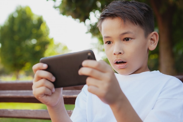 Junger asiatischer Junge, der Spiele an seinem intelligenten Telefon am Park spielt