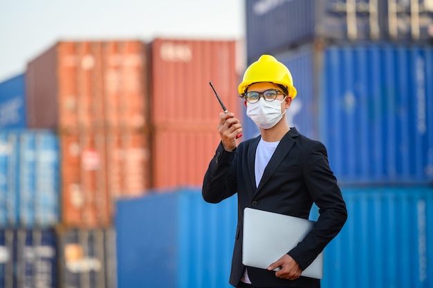 Junger asiatischer männlicher Ingenieur oder Manager Verwendung von Funkkommunikation und Laptops zur Steuerung der Containerladung in industriellen Transport- und Logistikkonzepten.