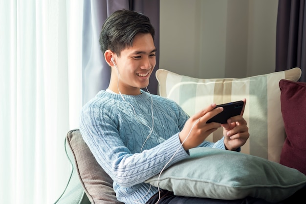 Junger asiatischer Mann, der auf dem Sofa liegt, Kopfhörer trägt und Telefon zu Hause spielt.