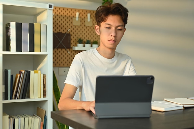 Junger asiatischer Mann, der auf ein digitales Tablet schaut, arbeitet oder recherchiert oder sich online auf eine Prüfung vorbereitet Bildung und Technologie