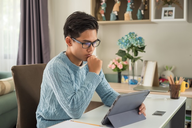 Junger asiatischer Mann, der Brille trägt, arbeitet an einer Tablette zu Hause.
