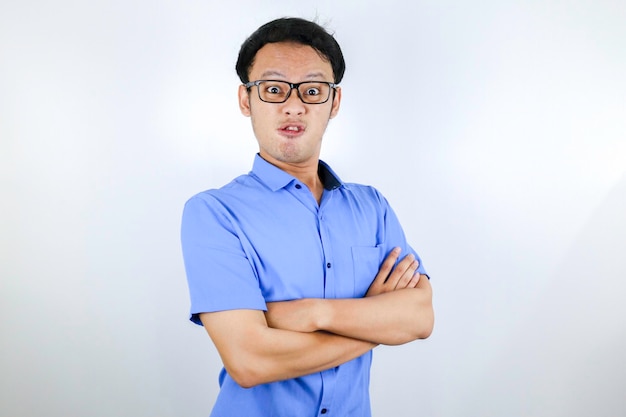 Junger asiatischer Mann, der ein blaues Hemd trägt, ist ein lustiges, wütendes Gesicht mit verschränkten Armen, das isoliert über weißem Hintergrund in die Kamera schaut