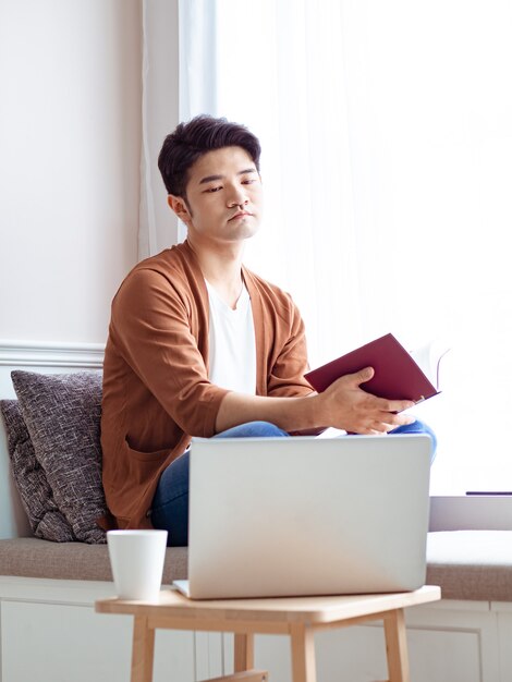 Junger asiatischer Mann, der ein Buch liest, während er sich zu Hause entspannt