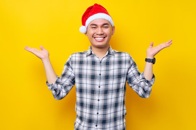 Junger asiatischer Mann, der ein kariertes Hemd in einer Weihnachtsmütze trägt, die Hände hebt und mit Aufregungsausdruck auf gelbem Hintergrund in die Kamera schaut Feier Weihnachtsfeiertag und Neujahrskonzept